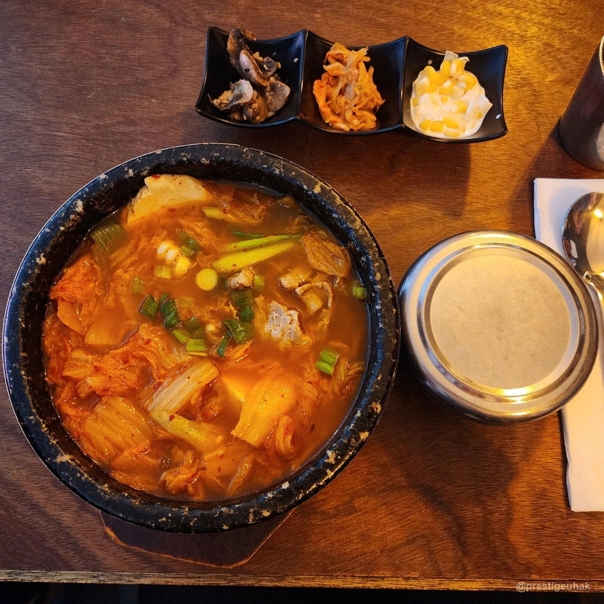 나무 식탁 위 검은 뚝배기에 담긴 붉은 찌개와 세 가지 반찬, 뚜껑 덮인 금속 밥그릇
