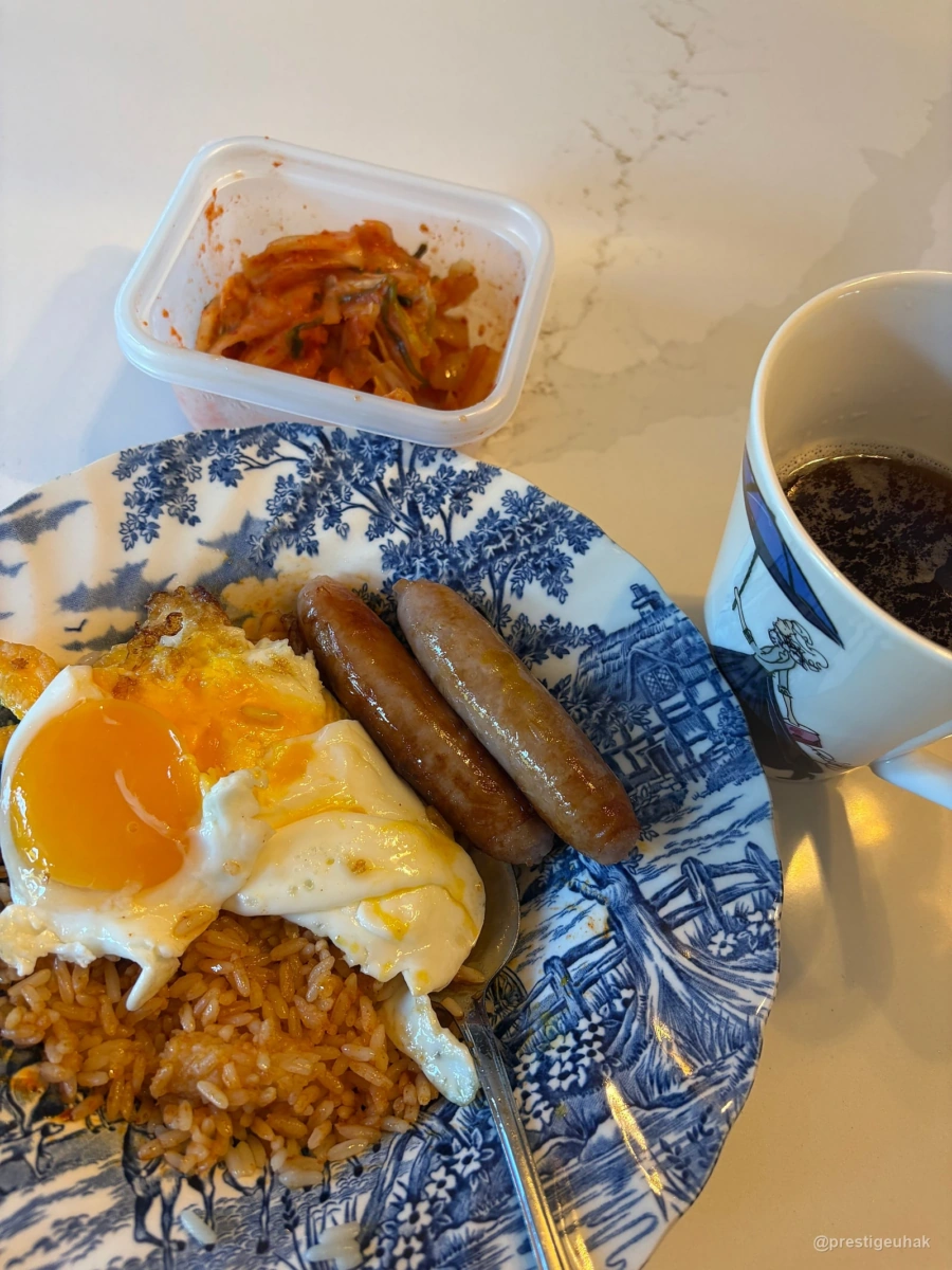 밥과 달걀프라이, 소시지가 담긴 파란 무늬 접시 옆에 김치 용기와 음료가 든 머그잔이 놓여 있다.